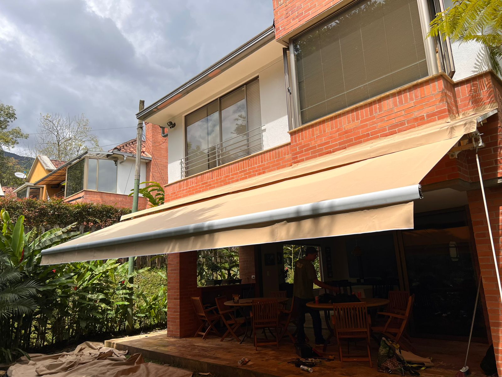 Baneta enrollable beige extendida sobre terraza de casa de ladrillo con jardín tropical